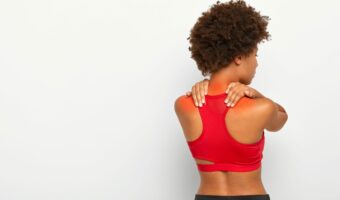 Back view of young curly woman suffers from neck pain and osteoporosis, has painful feelings in muscles, holds hands near shoulders, wears red top, isolated over white background. Health issues