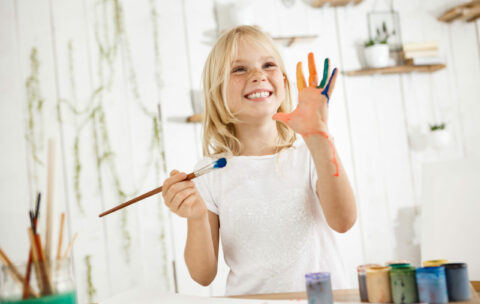 Happy and playful cute freckled blonde girl dressed in white, holding brush in one hand and showing another hand, which she messed up with paint. Children and happiness concept. Isolated shot, horizontal