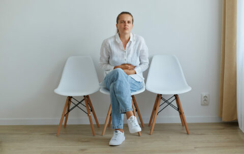Portrait of sick woman with dark hair and ponytail, wearing white shirt and jeans, sitting on chair in queue to the doctor in clinic, suffering from terrible stomachache.